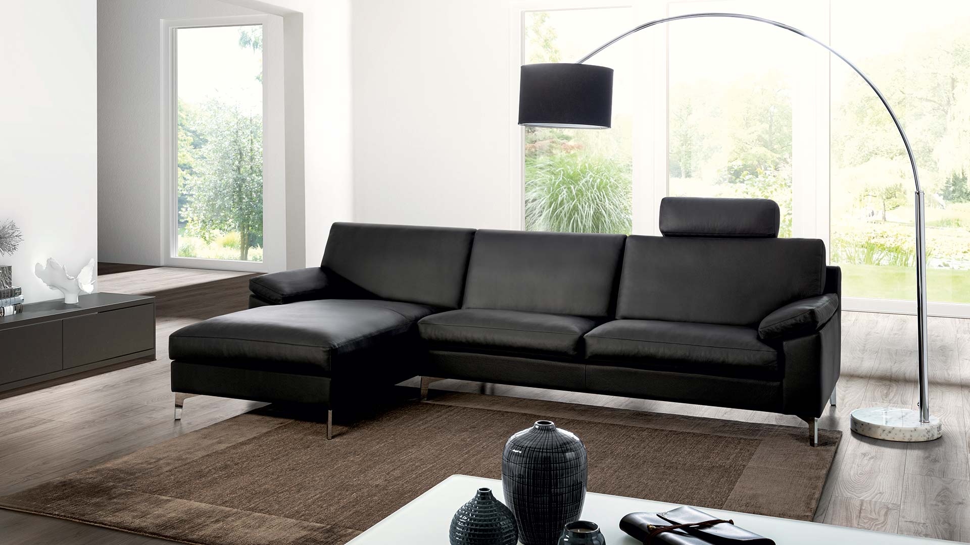 Helles Wohnzimmer mit schwarzem Erpo Ledersofa als Longchair mit Kopfstütze und silbernen Metallfüßen.