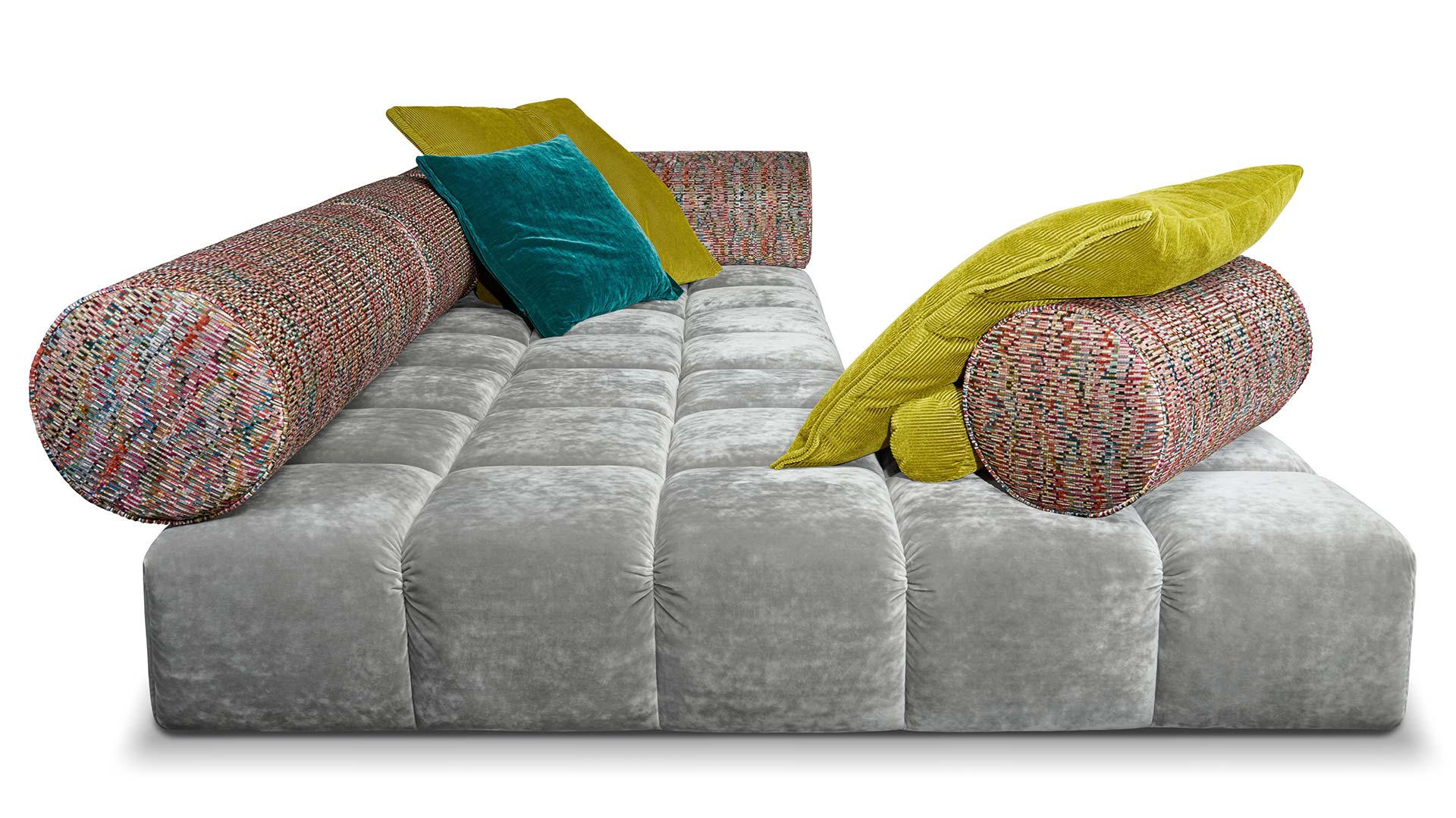 Bodentiefe Ed Couch von Bretz silbernem Velours und bunt gemusterten Polsterrollen und blauem und gelbem Plaidkissen in Velours und Cord.