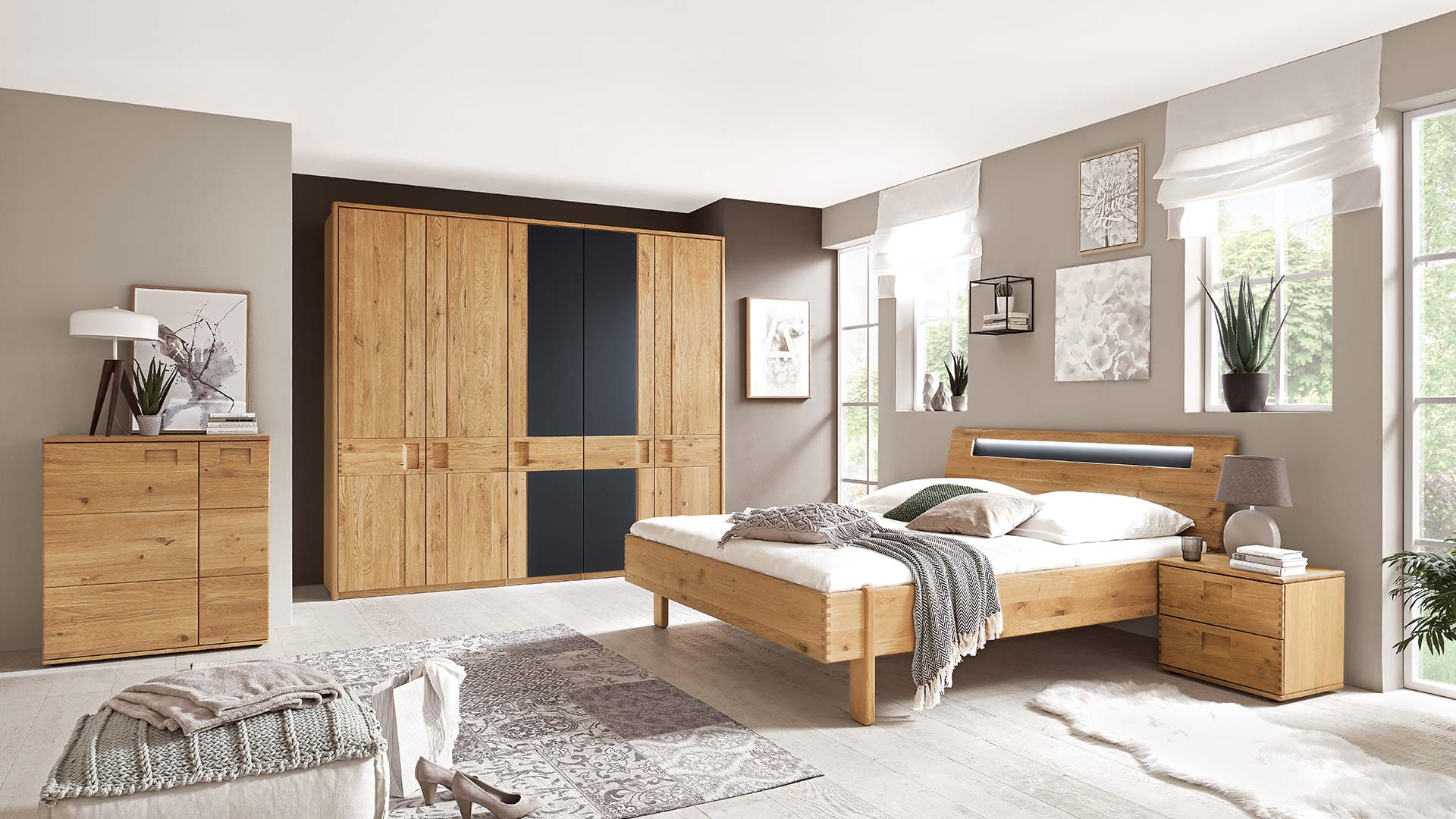 Schlafzimmersystem in einem beigen Schlafzimmer mit Massivholzbett in Eiche oder Buche bleuchtet mit Nachtkonsole, Kleiderschrank mit schwarzen Akzenten und Kommode.