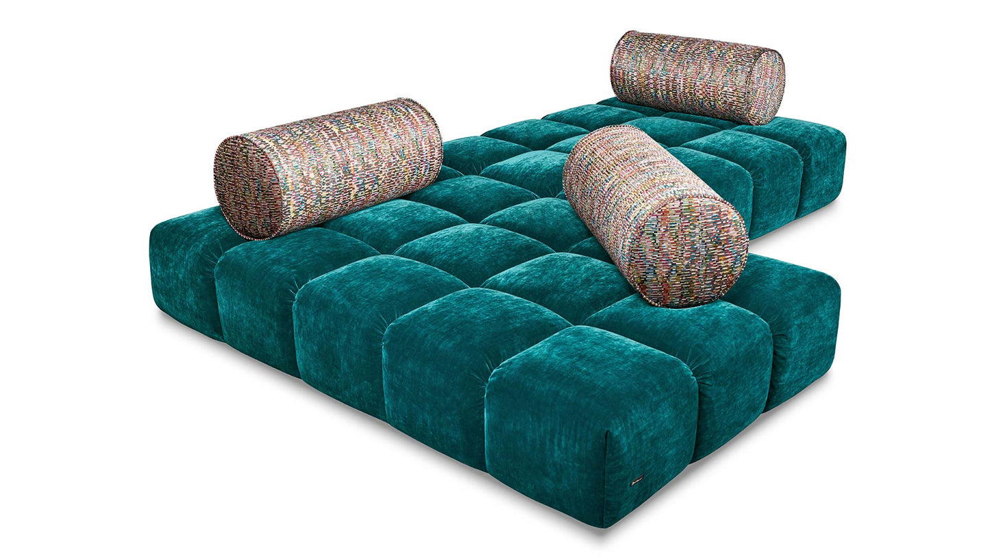 Bodentiefe Ed Couch von Bretz pertrolfarbendem Velours und bunt gemusterten Polsterrollen.